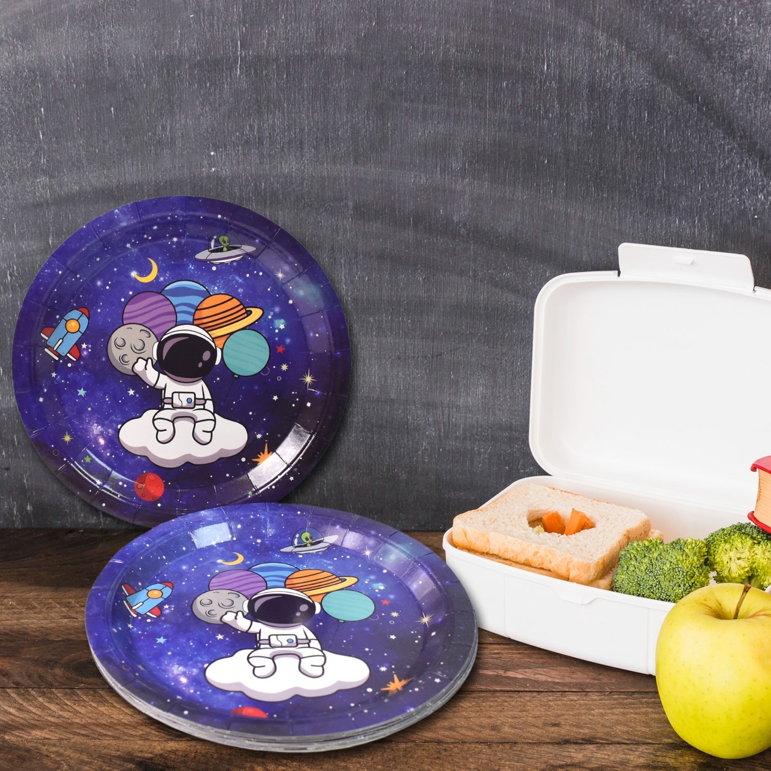 Two purple paper plates with an astronaut and planets design, next to an open lunchbox and an apple.