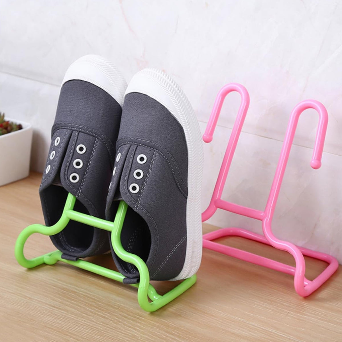 A pair of grey canvas shoes hangs from a green plastic drying rack next to a pink rack on a wooden surface.