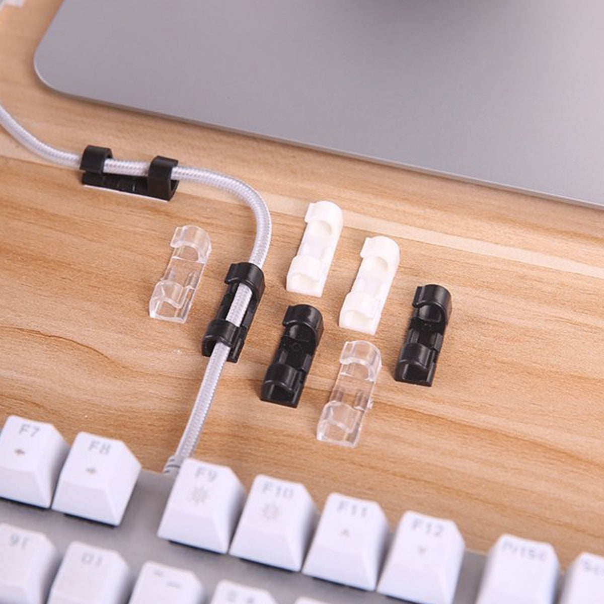 A mix of black, white, and clear plastic cable clips on a wooden desk next to a white keyboard and laptop.