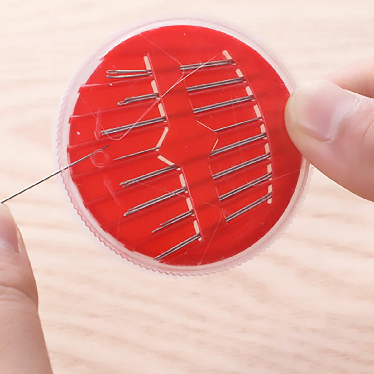 Close-up of hands holding a red disc containing multiple sewing needles, with one needle being removed.