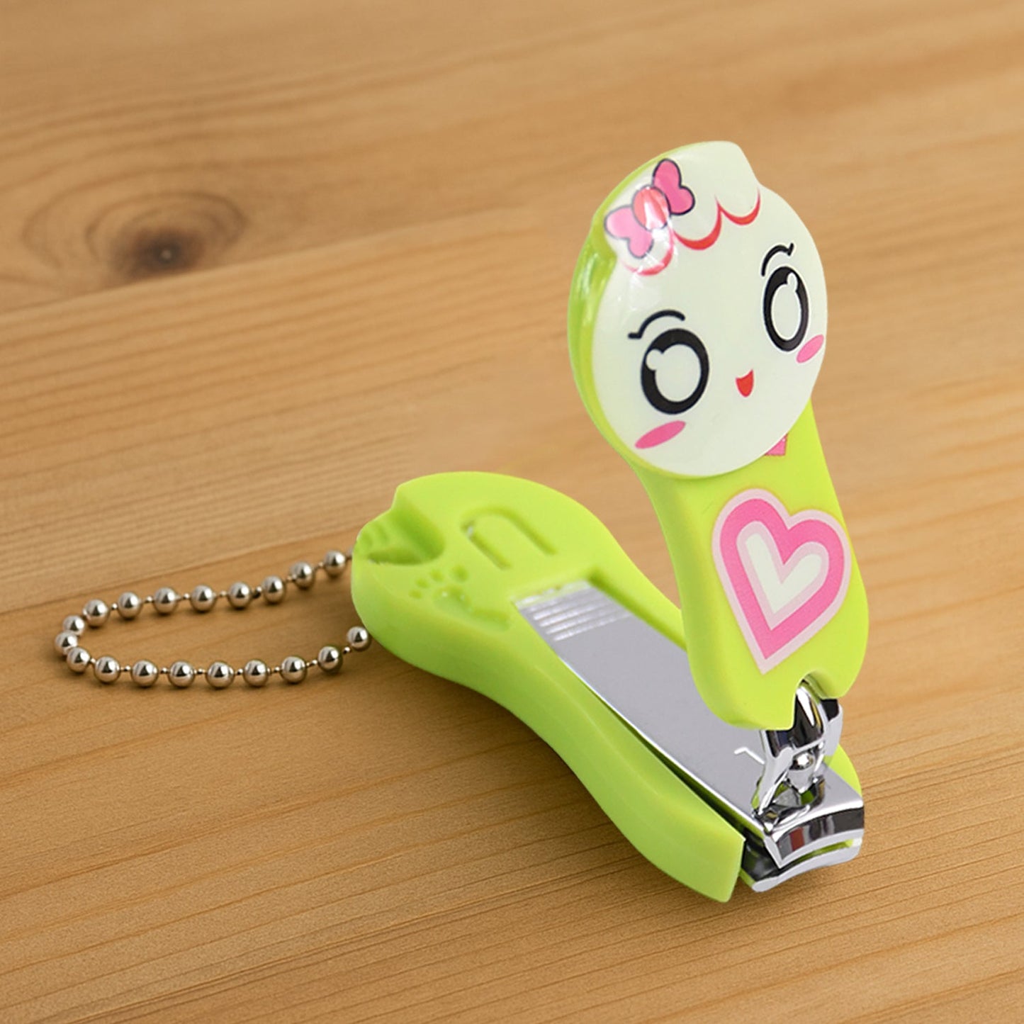A green and white cartoon character nail clipper with a pink heart and a silver ball chain, on a wooden surface.