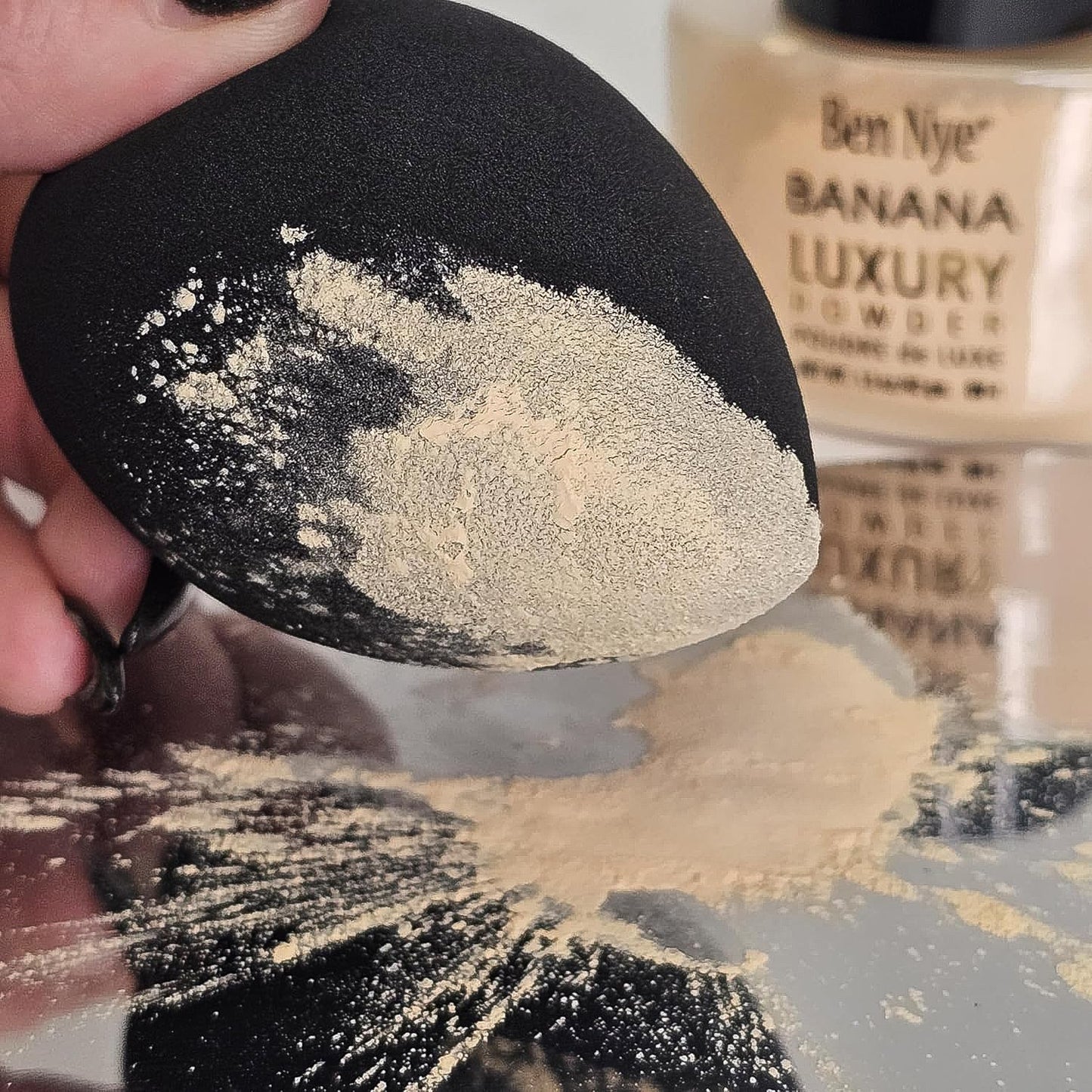 A black makeup sponge covered in light beige setting powder, with a Ben Nye Banana Luxury Powder jar in the background.
