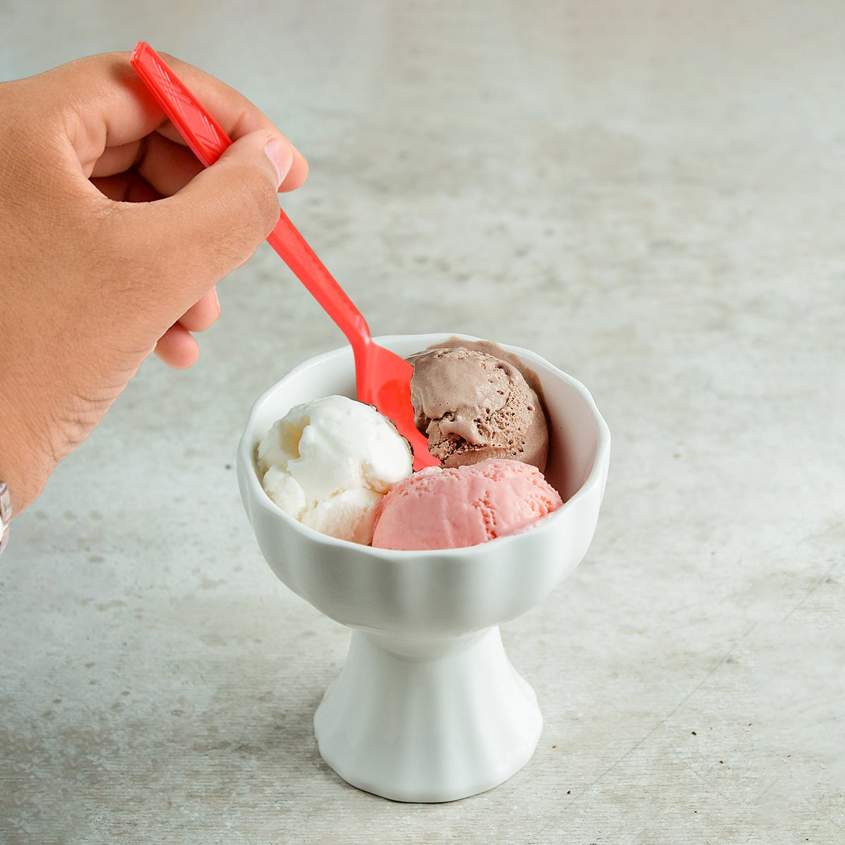 A hand holding a red plastic spoon scooping ice cream from a white bowl containing three flavors.