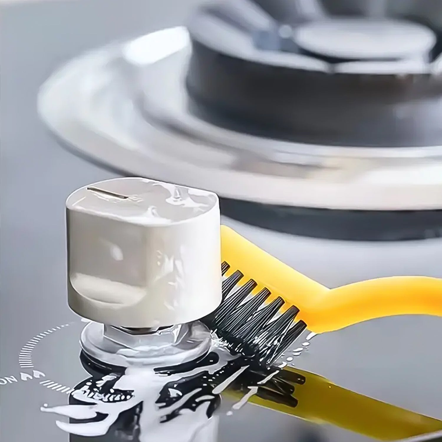 A yellow wire brush cleans around a gas stove burner knob on a black stovetop, with suds visible.