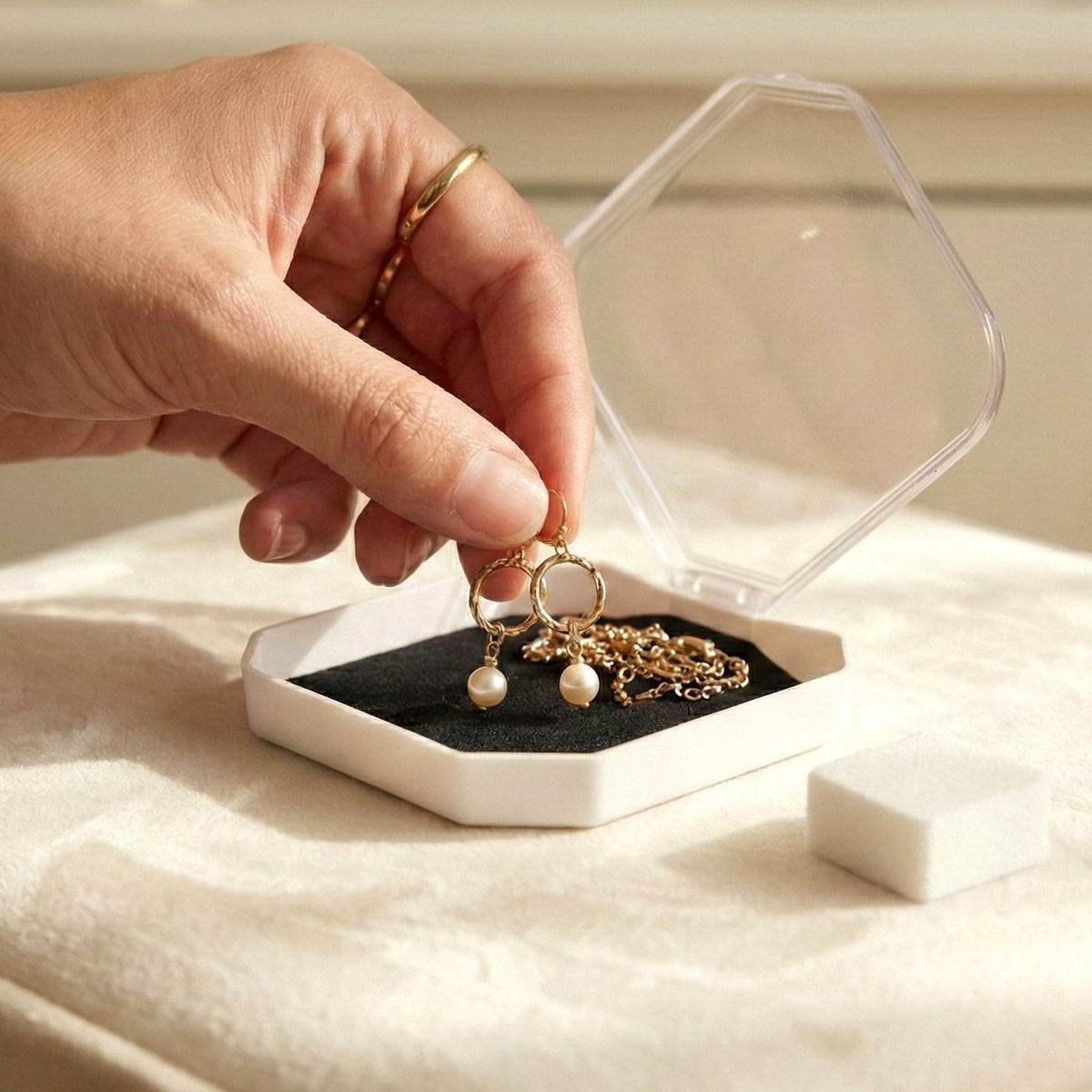 A hand holding gold earrings with pearls over an open jewellery box with a gold necklace inside.