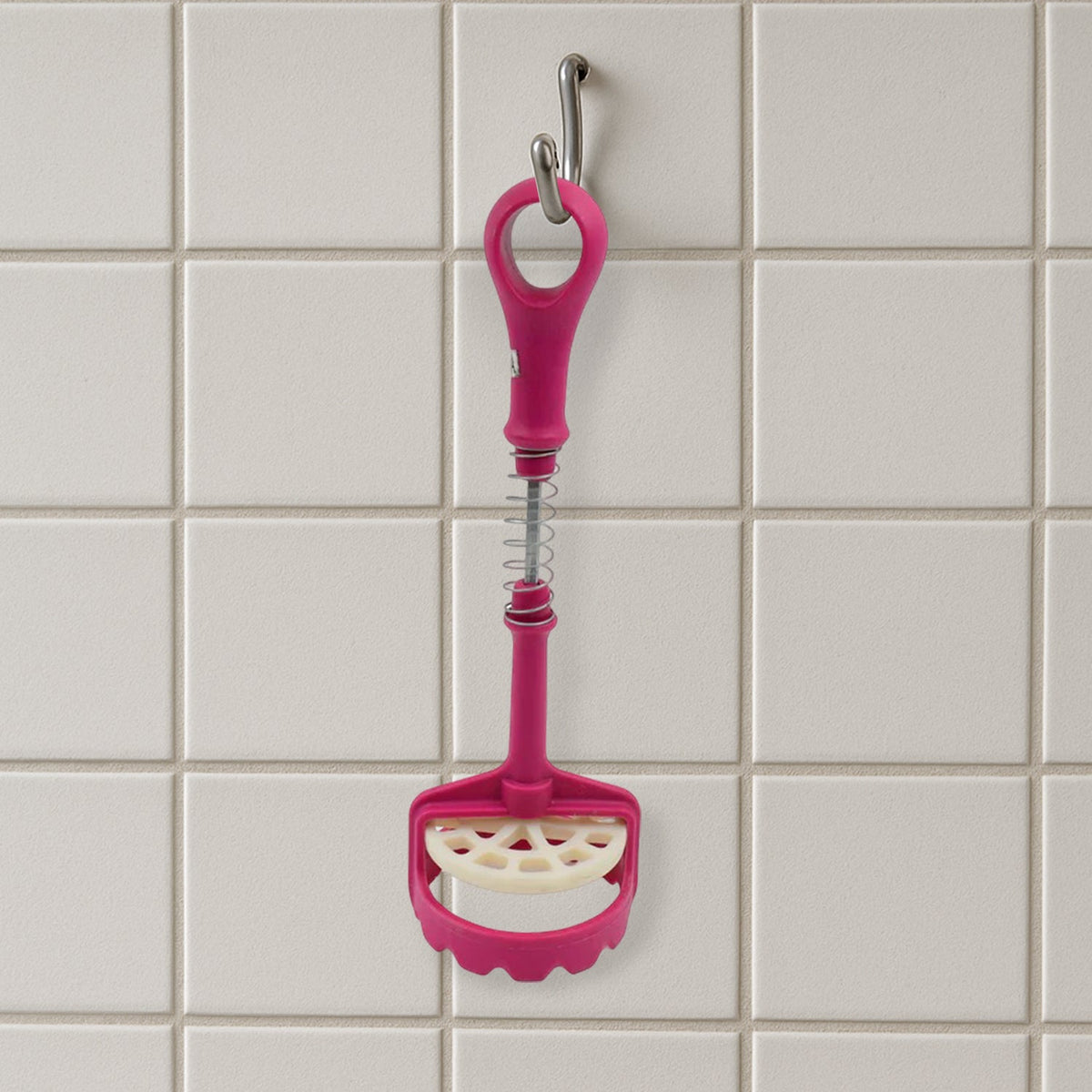 A pink and white plastic potato masher hangs vertically from a silver hook against a white tiled wall.