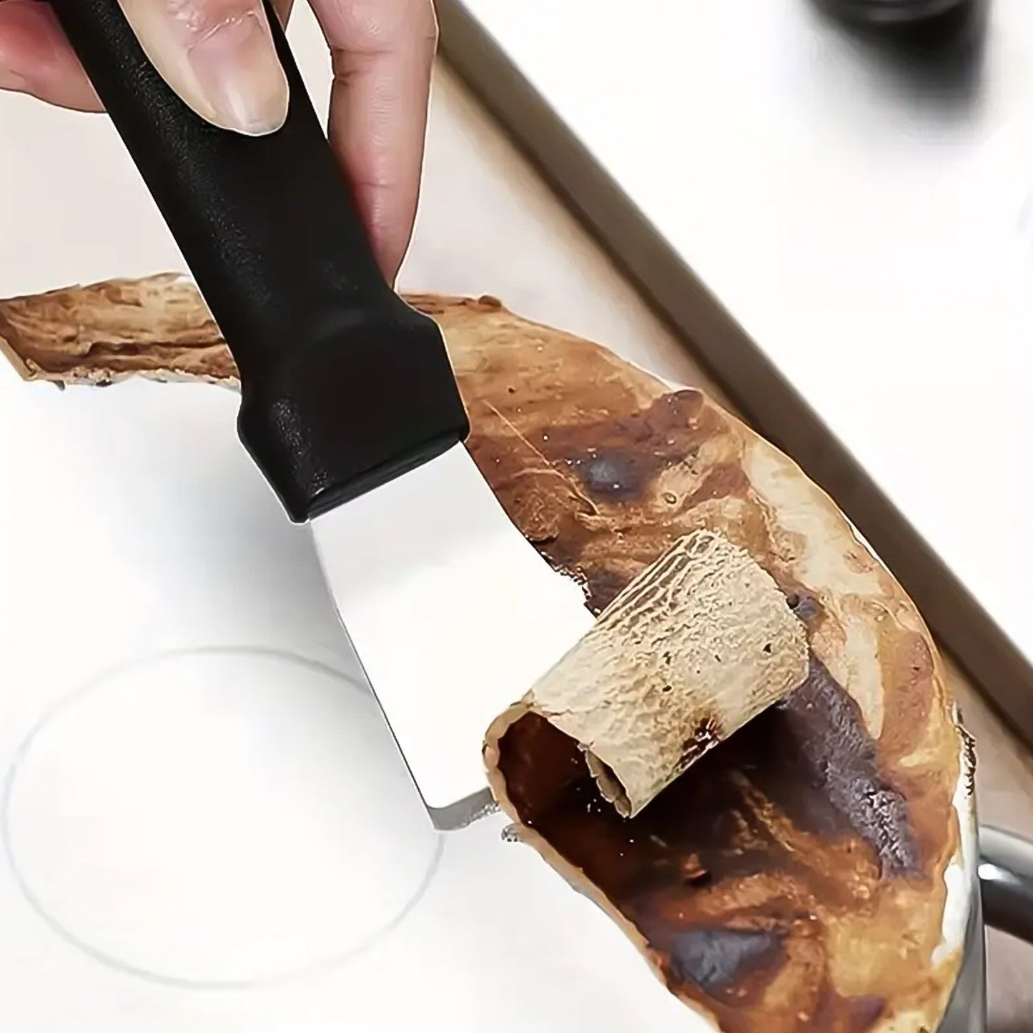 A hand uses a black scraper to remove burnt residue from a white surface, peeling it away in strips.
