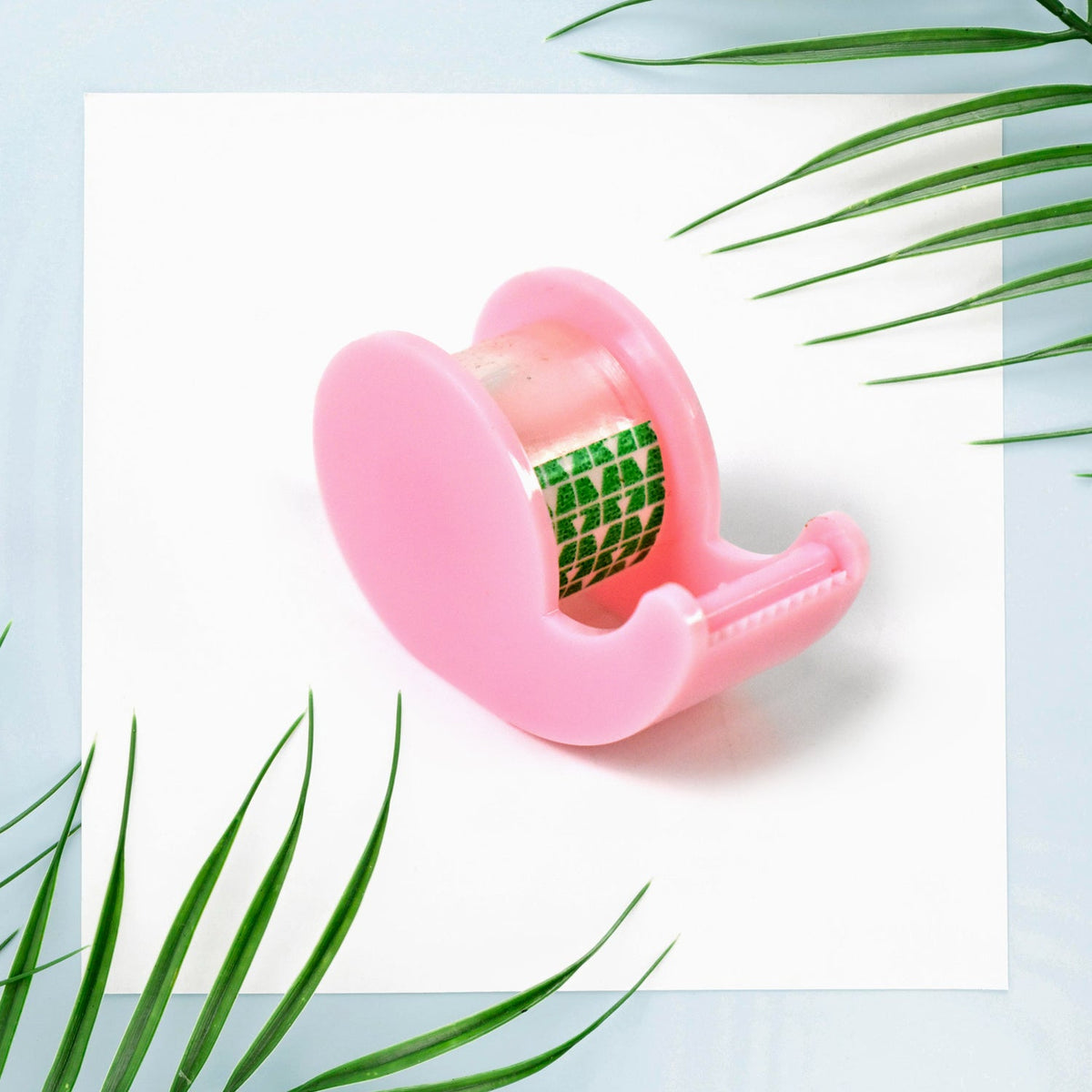 A pink tape dispenser with a roll of clear tape, featuring green patterns, on a white square with palm leaves.