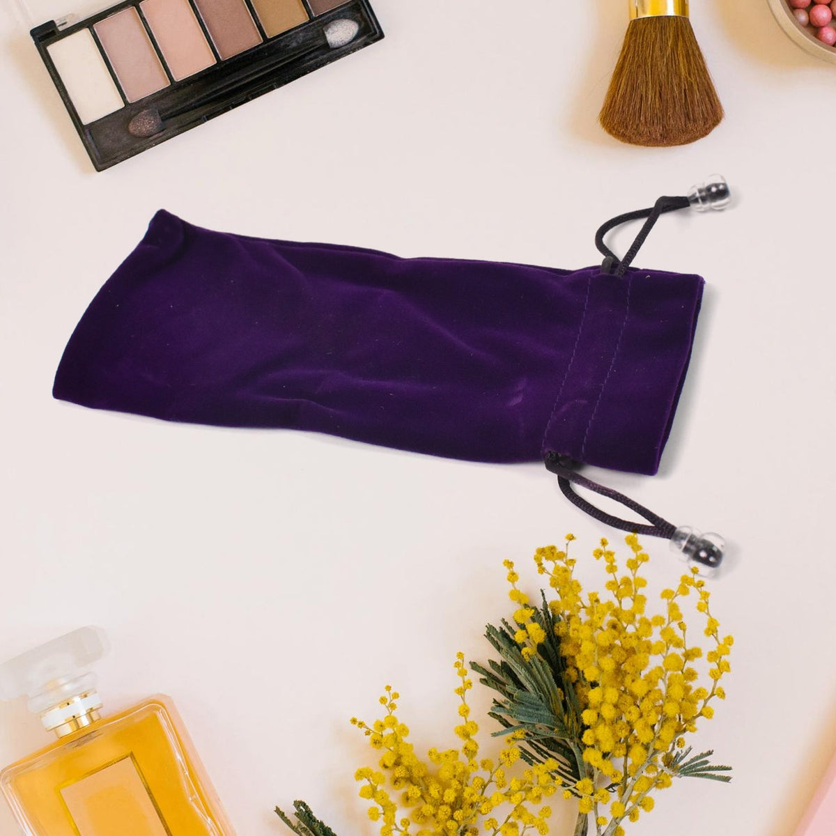 A purple velvet drawstring bag with a DayShow logo is on a white surface surrounded by makeup, perfume, and yellow flowers.