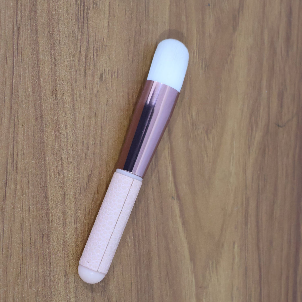 A pink and rose gold makeup brush with a white brush head, angled on a wooden surface.