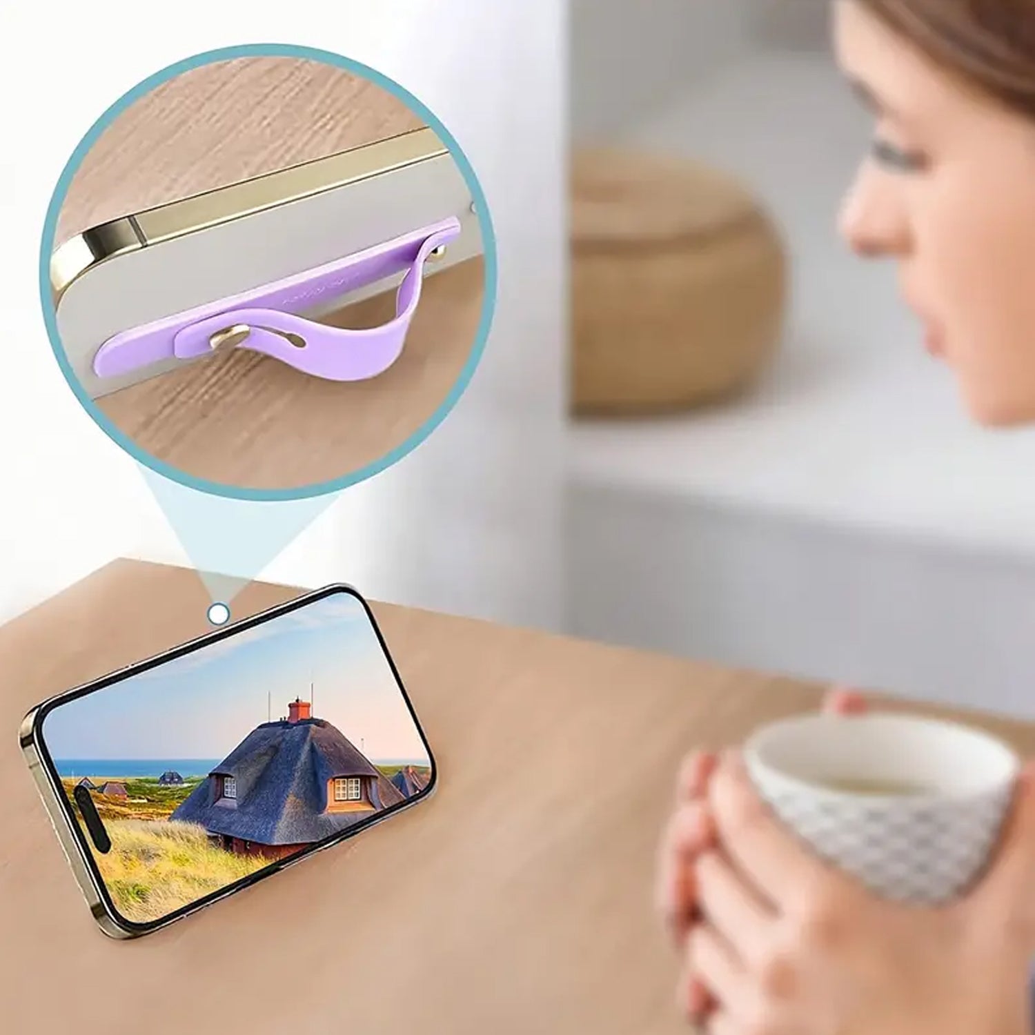 A phone propped up by a purple silicone grip, showing a landscape image, with a woman holding a cup in the background.