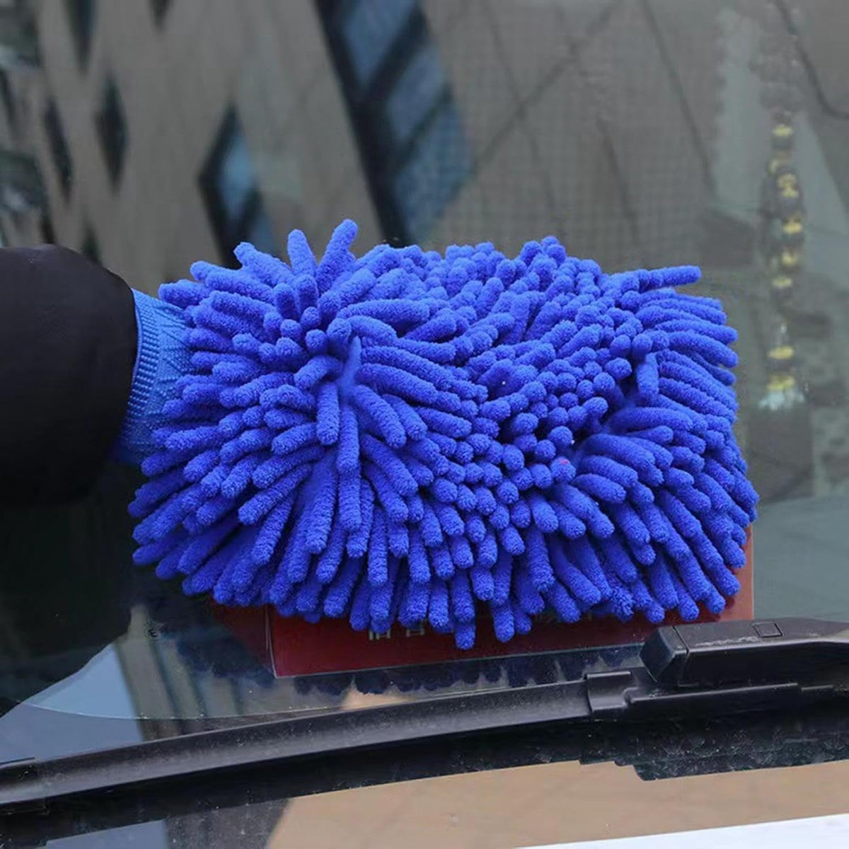A blue microfiber wash mitt held by a hand, cleaning the windshield of a car.