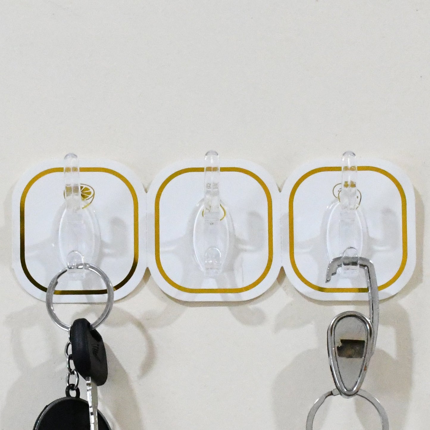 Three transparent adhesive hooks with gold borders on a white wall, holding two keychains.
