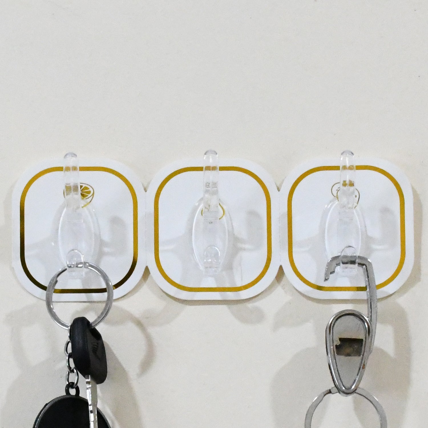 Three transparent adhesive hooks with gold borders on a white wall, holding two keychains.