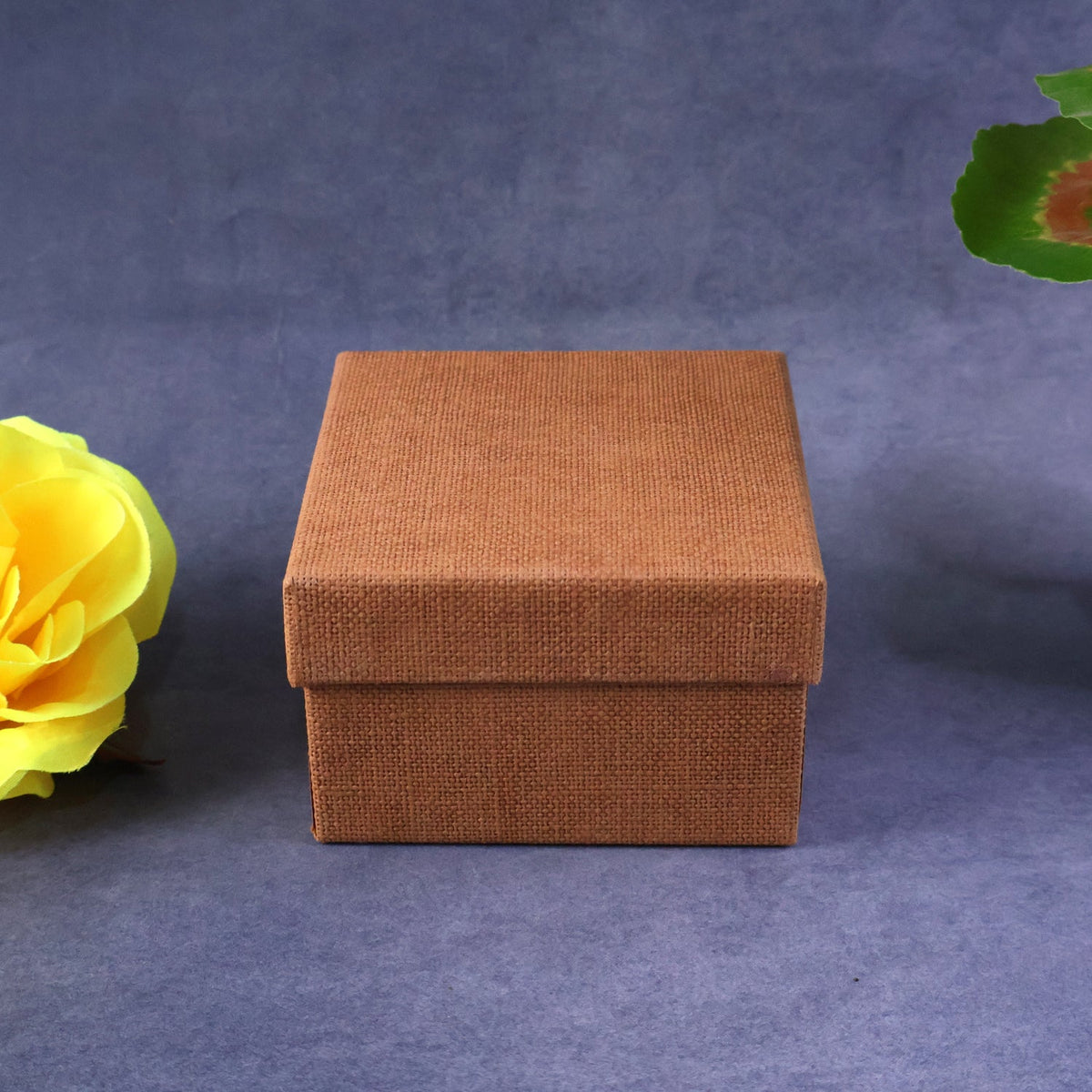 A closed, square, brown textured gift box on a dark grey background with a yellow flower.