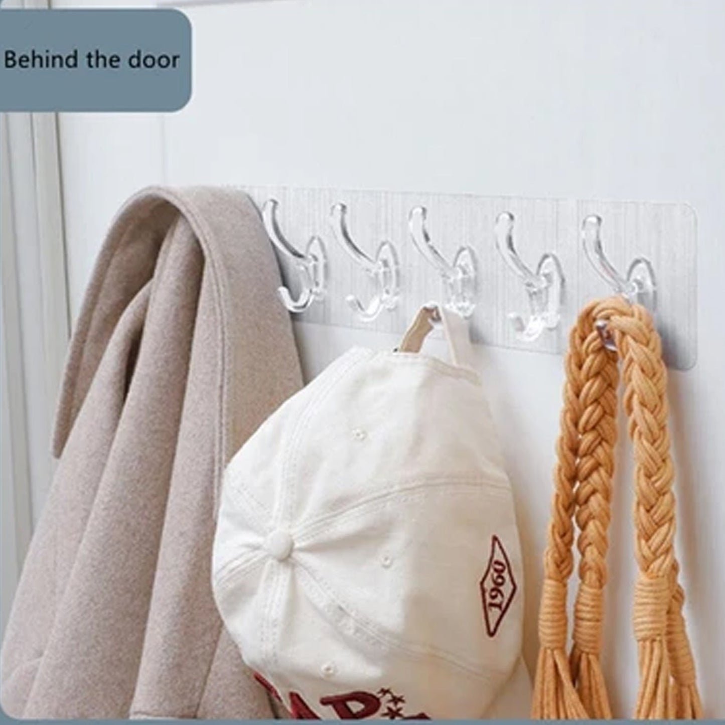 A clear 6-hook adhesive rail holds a beige coat, white baseball cap, and braided strap on a white door.