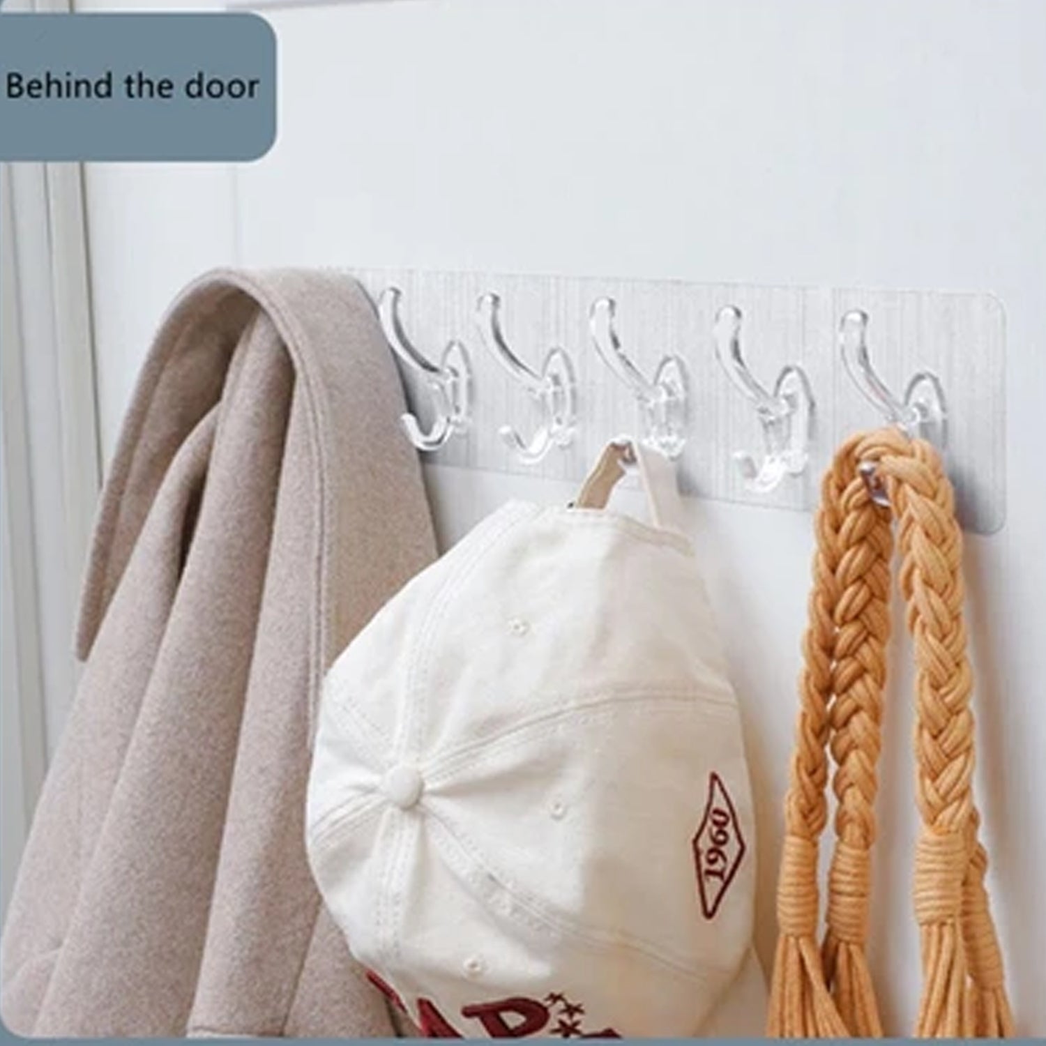 A clear 6-hook adhesive rail holds a beige coat, white baseball cap, and braided strap on a white door.