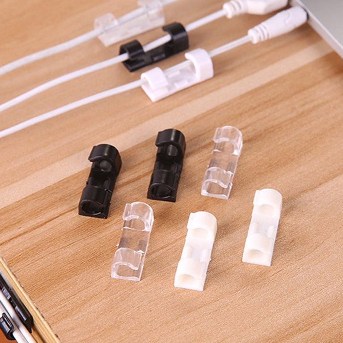 Six cable clips in black, clear, and white plastic on a wooden surface, with cables secured by other clips in the background.