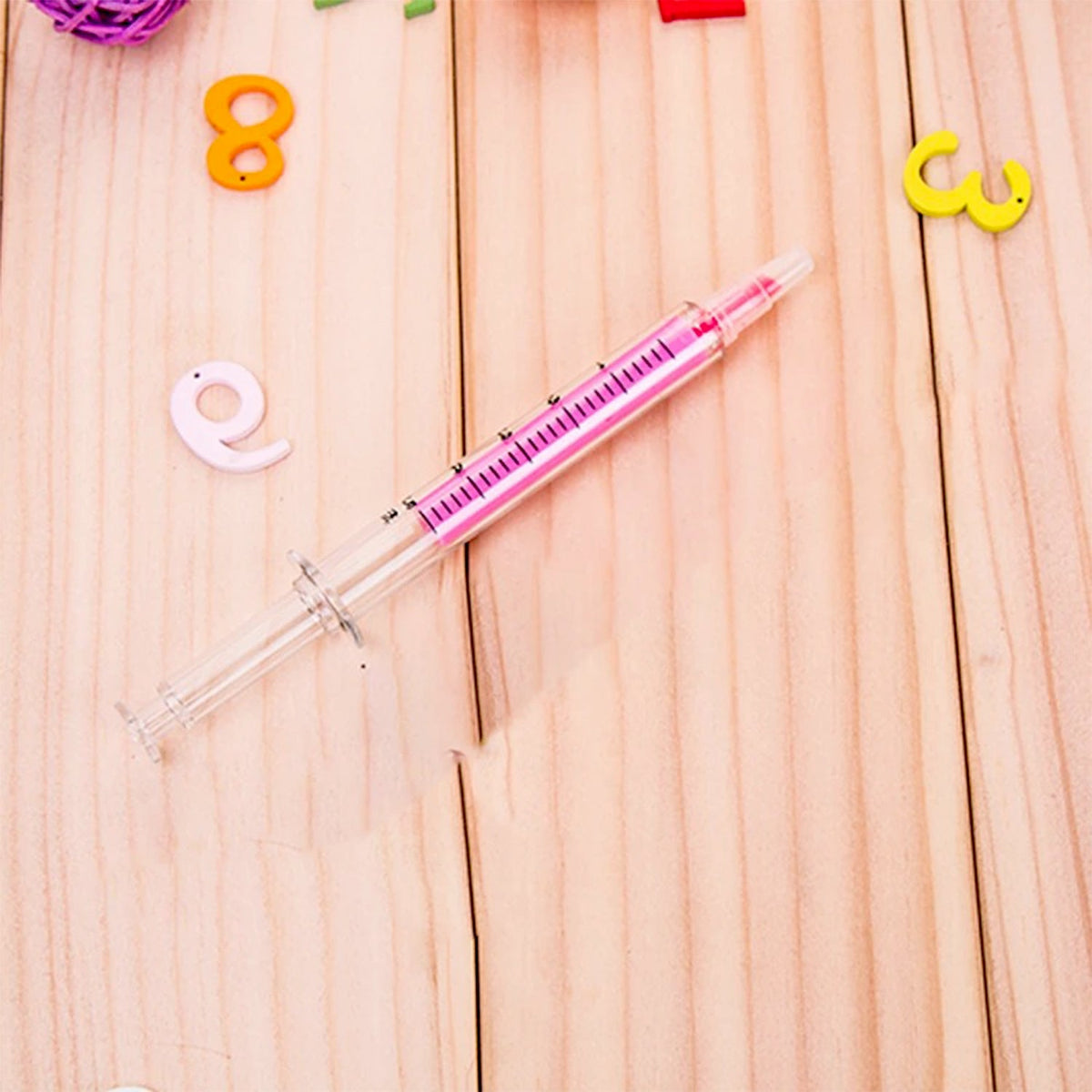A pink syringe highlighter with a clear barrel and measurements, lying on a light wooden surface with colorful numbers.