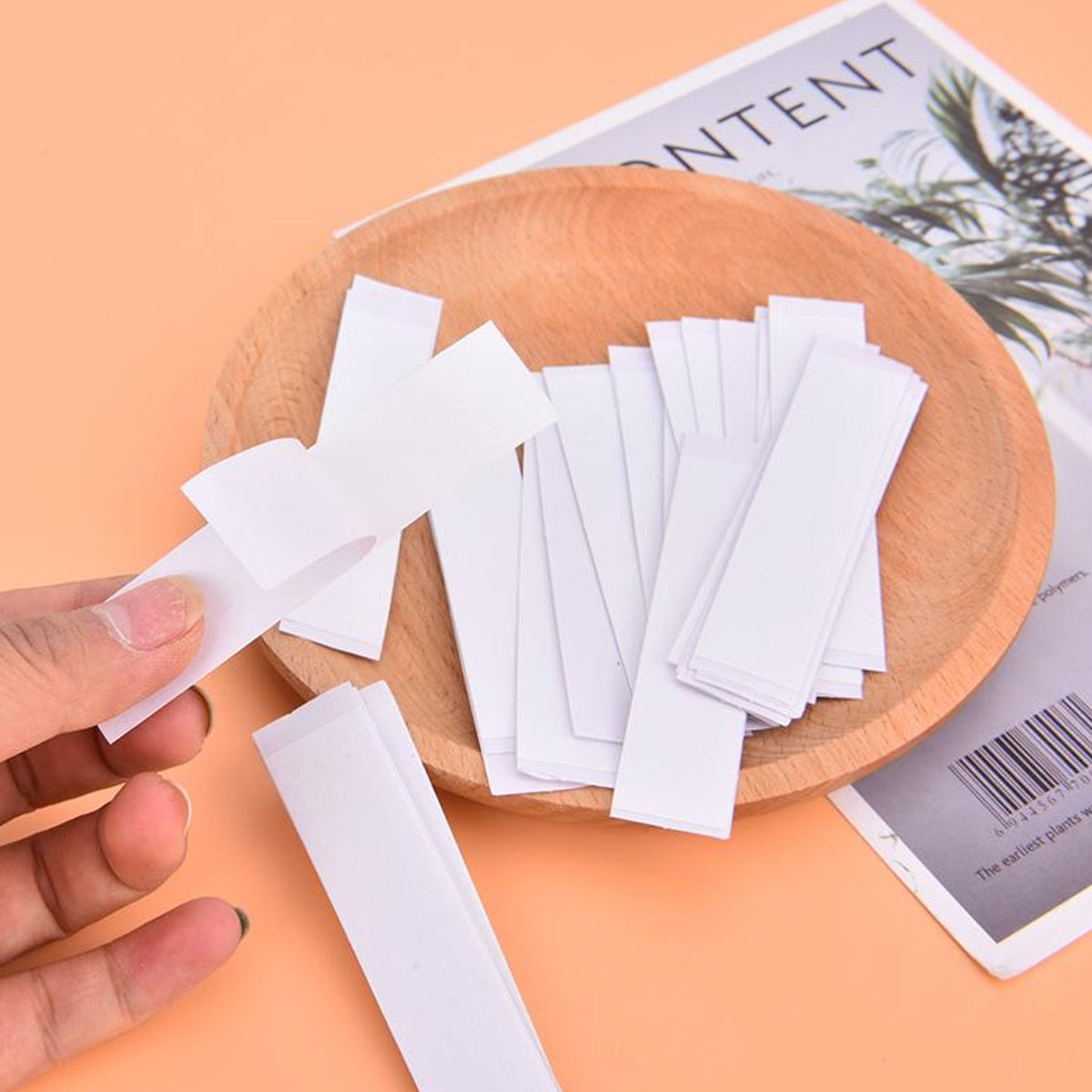 A hand holds a white strip of tape over a wooden bowl filled with many similar strips, next to a magazine.
