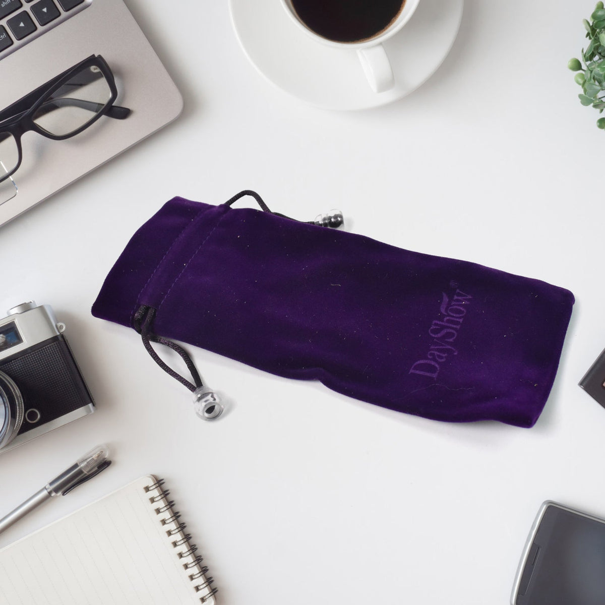 A purple velvet drawstring bag with a DayShow logo is on a white desk with a laptop, coffee, and glasses.