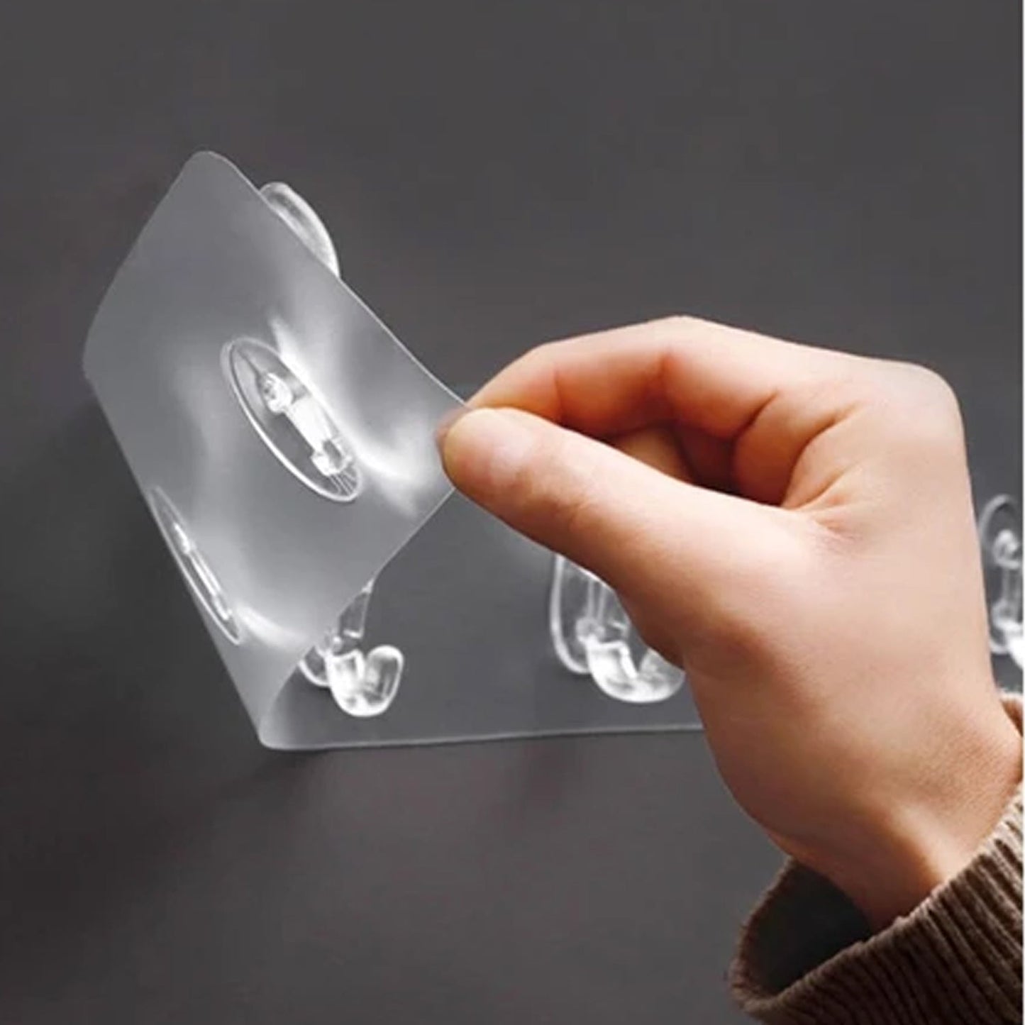 A hand peels back a corner of the clear plastic adhesive wall hook rail from a dark surface.