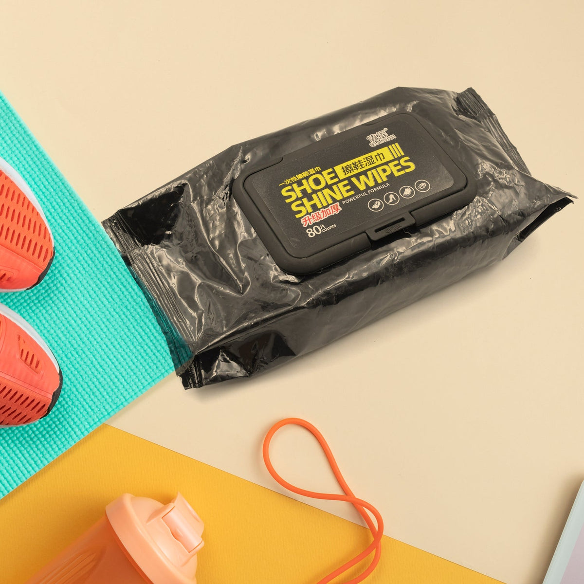 A black pack of "Shoe Shine Wipes" rests on a teal mat next to an orange shoe and water bottle.