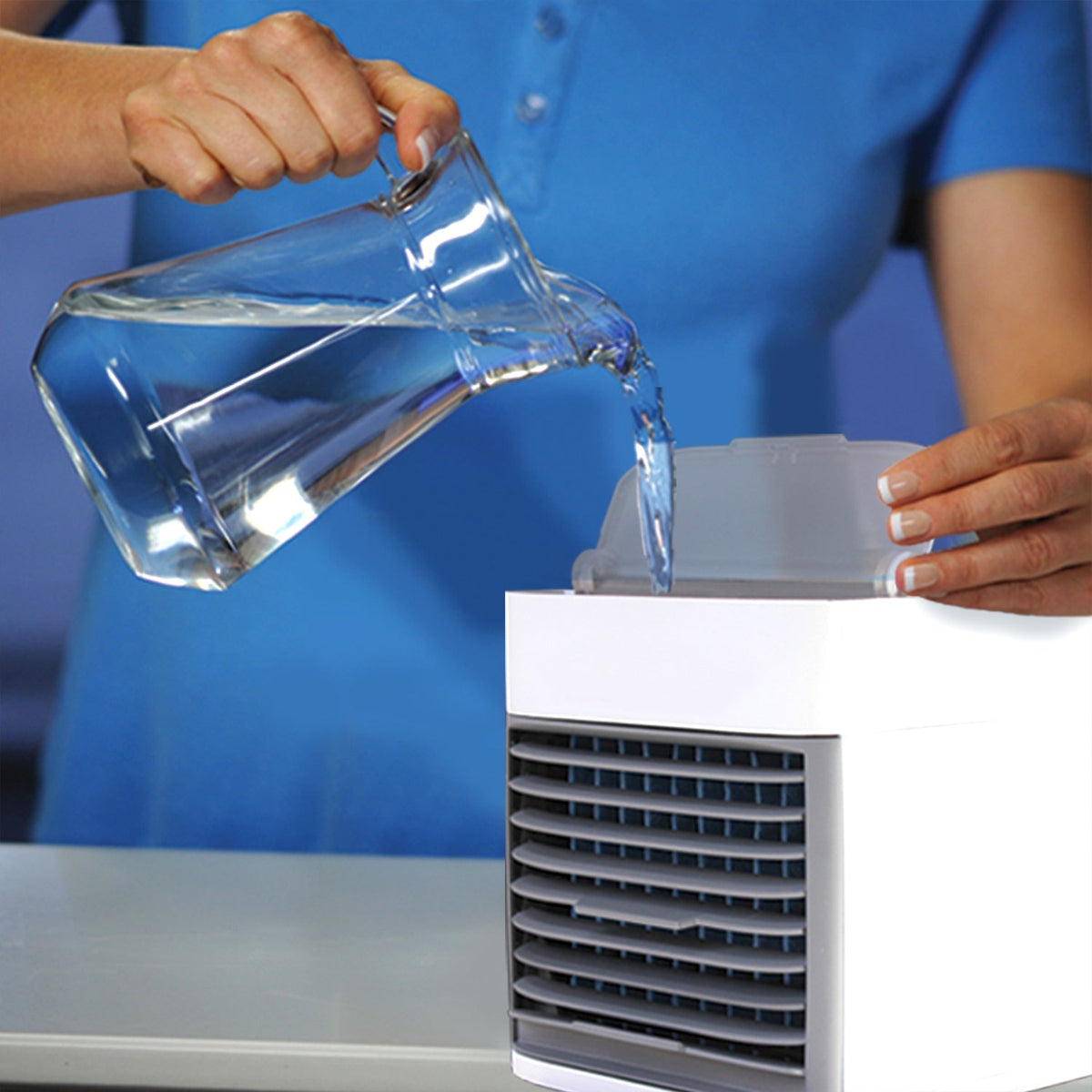 Hands pouring water from a clear pitcher into the top reservoir of a white and gray portable air cooler.