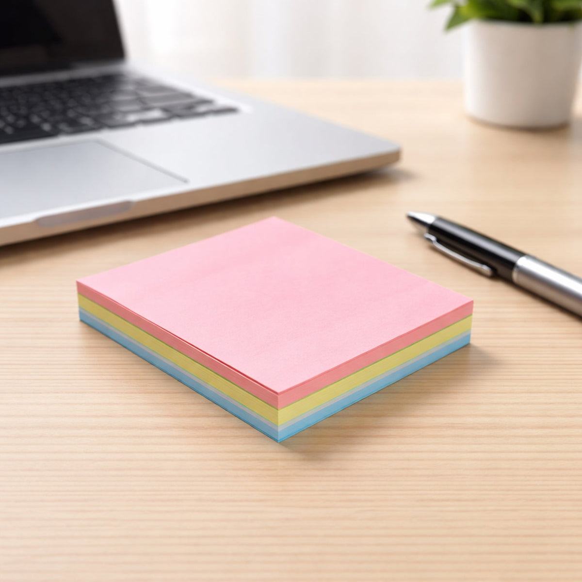 A stack of pink, yellow, and blue sticky notes on a wooden desk, with a silver laptop and a black pen in the background.