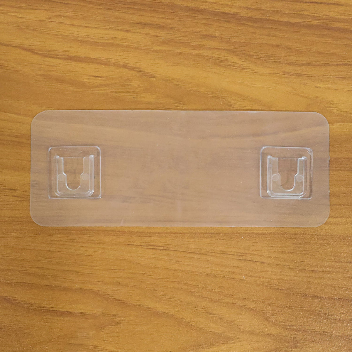 A clear plastic self-adhesive hanger with two hooks is placed on a light brown wooden surface.