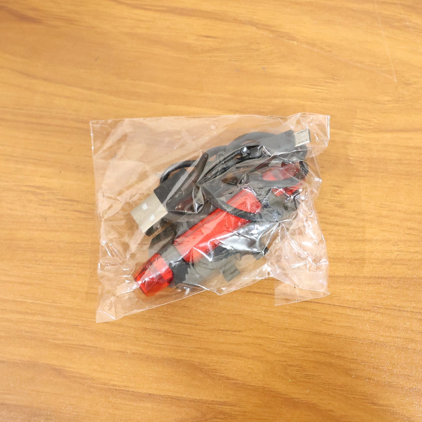 A clear plastic bag containing a red LED bicycle tail light and its accessories rests on a light brown wooden surface.