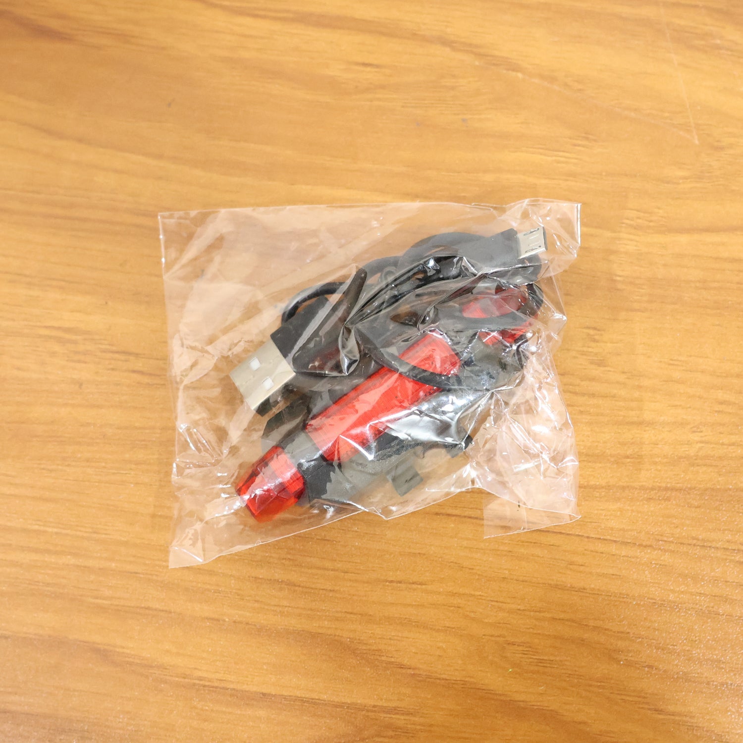 A clear plastic bag containing a red LED bicycle tail light and its accessories rests on a light brown wooden surface.