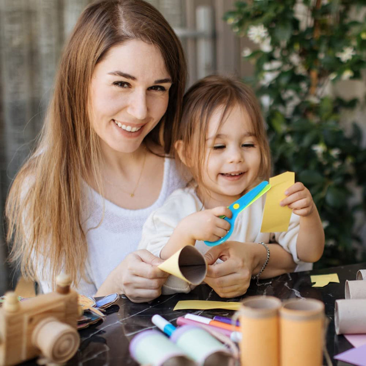 A smiling woman and a young child craft together, the child cutting yellow paper with blue and yellow scissors.