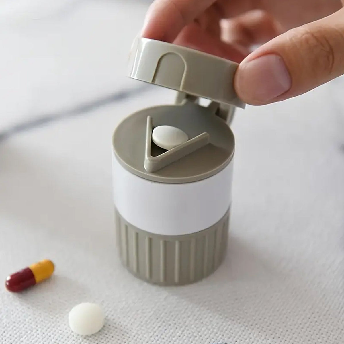 A hand opening a gray and white pill cutter with a white pill inside and a capsule nearby.