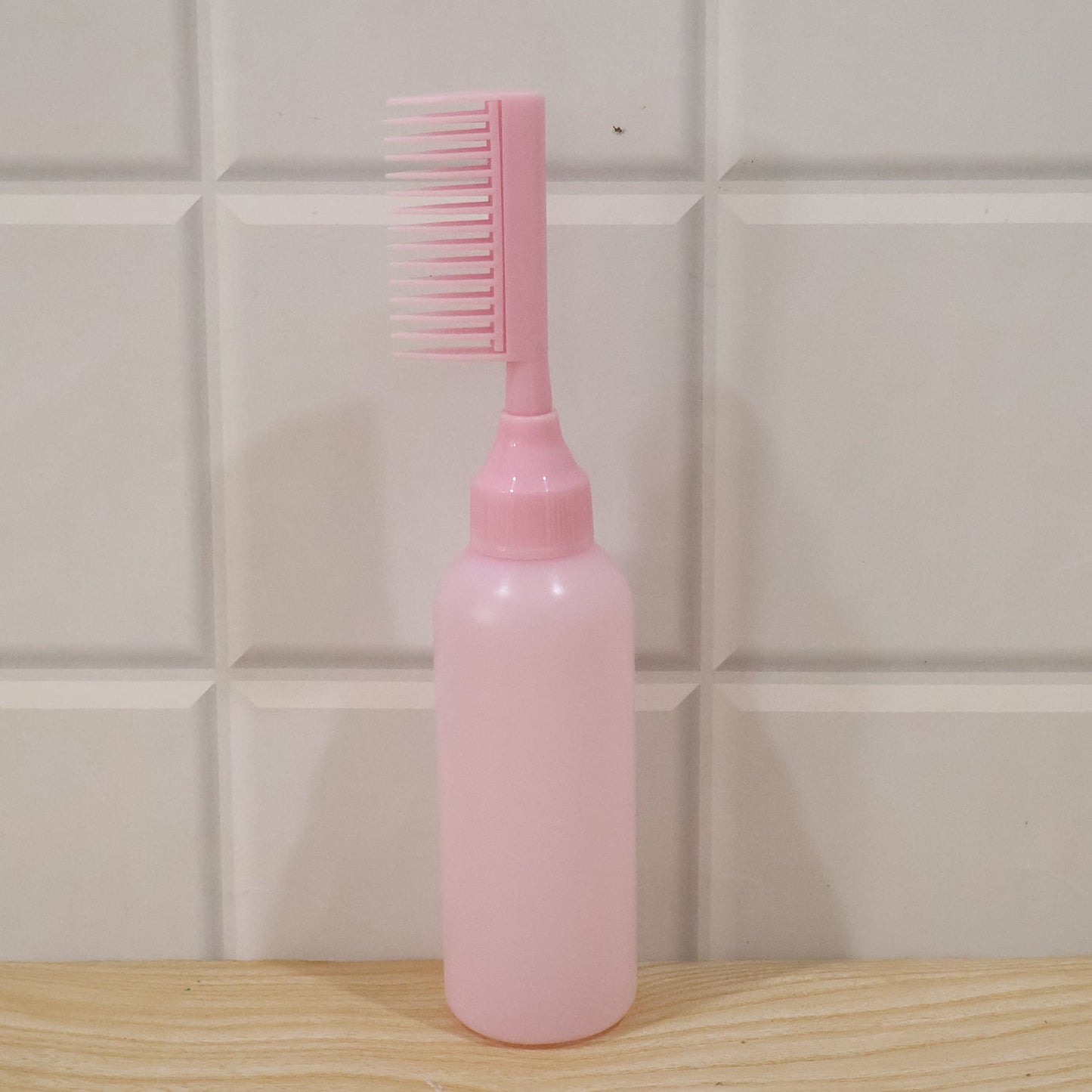 A pink 2 In 1 Hair Oil Applicator Bottle with Scalp Comb stands on a wooden surface against a white tiled wall.