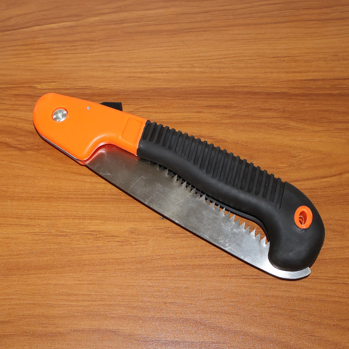 The orange and black folding hand saw, with its metal blade folded into the handle, rests on a wooden surface.