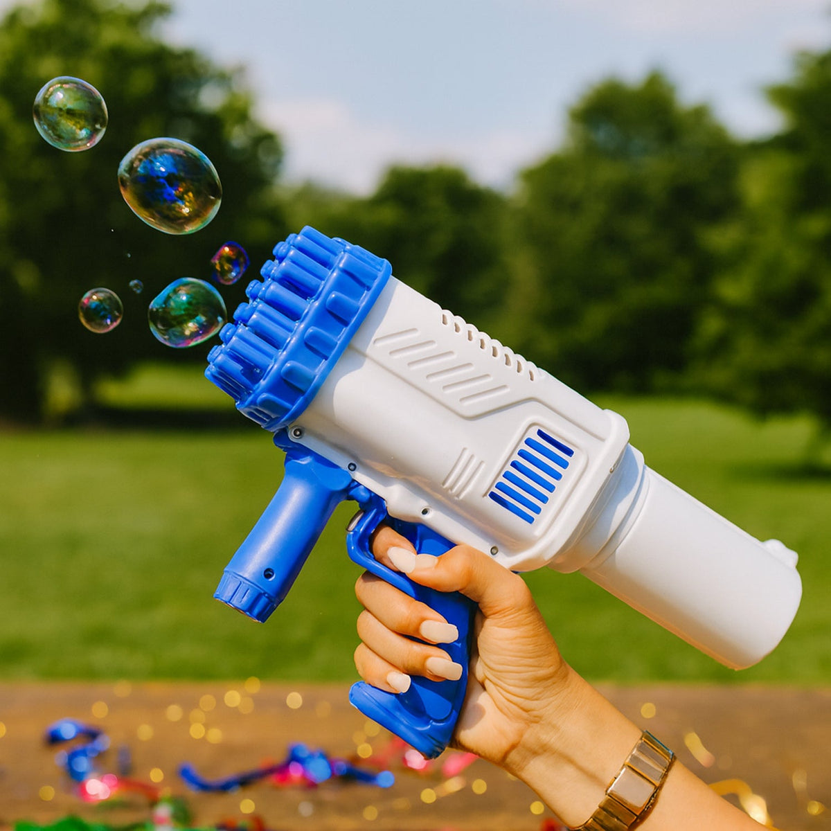A hand holds the white and blue 32-hole bubble gun, blowing bubbles outdoors with green trees in the background.