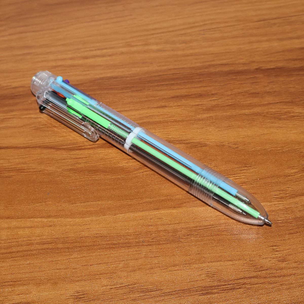 A clear 6-in-1 multicolor ballpoint pen with blue and green ink cartridges is angled on a warm brown wooden surface.