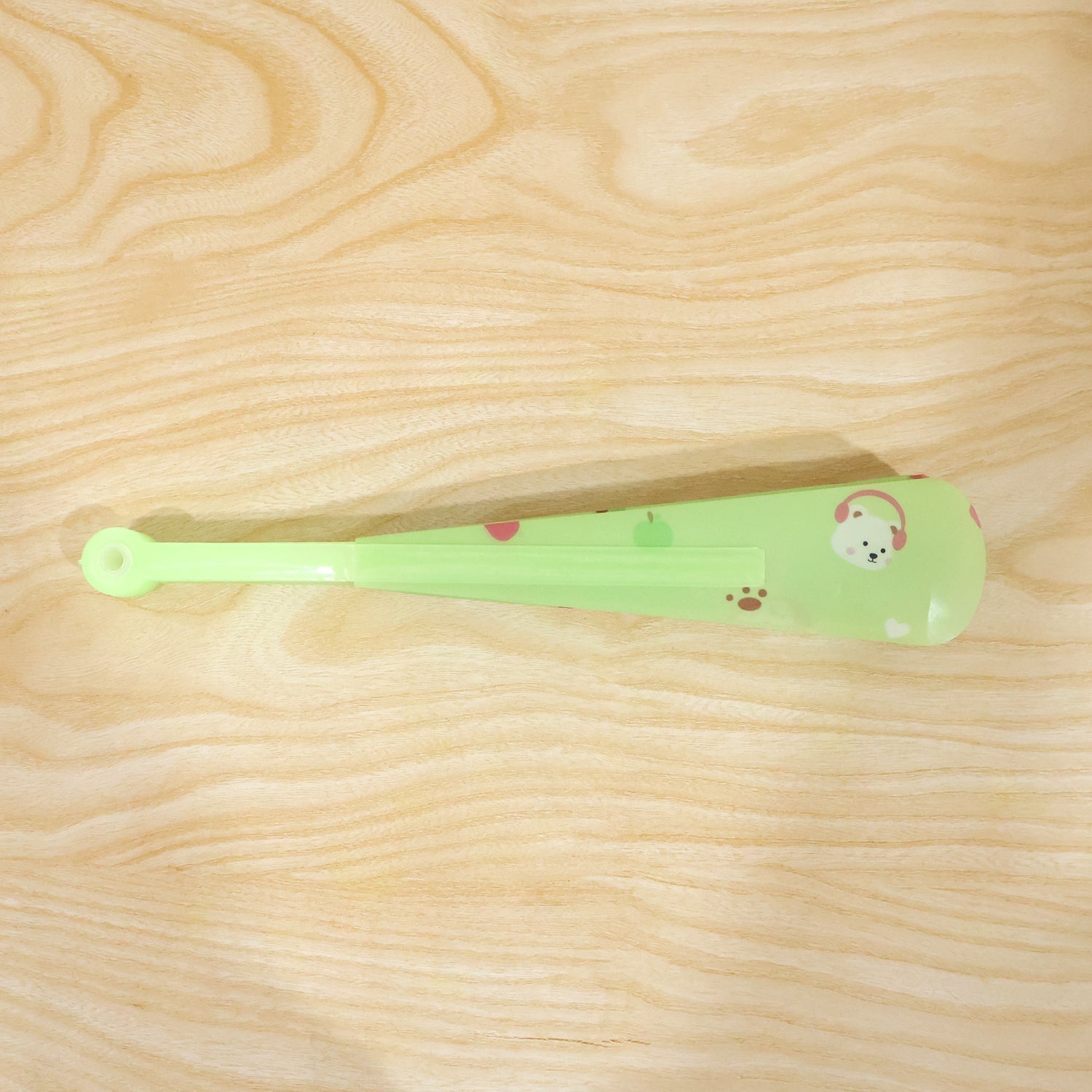 A folded green plastic fan with a bear and apple pattern lies on a light wooden surface.