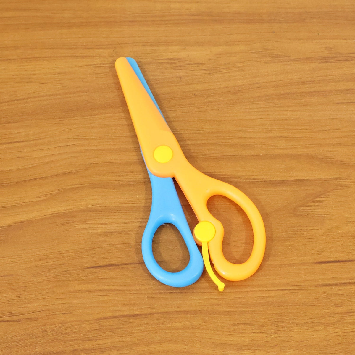 A pair of blue and orange plastic scissors with yellow accents rests closed on a light brown wooden surface.