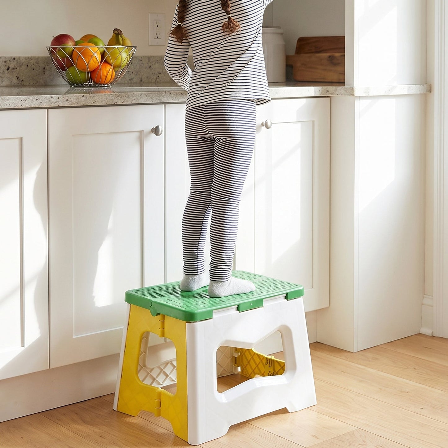 A child stands on a green, white, and yellow plastic stool to reach items on a kitchen counter.
