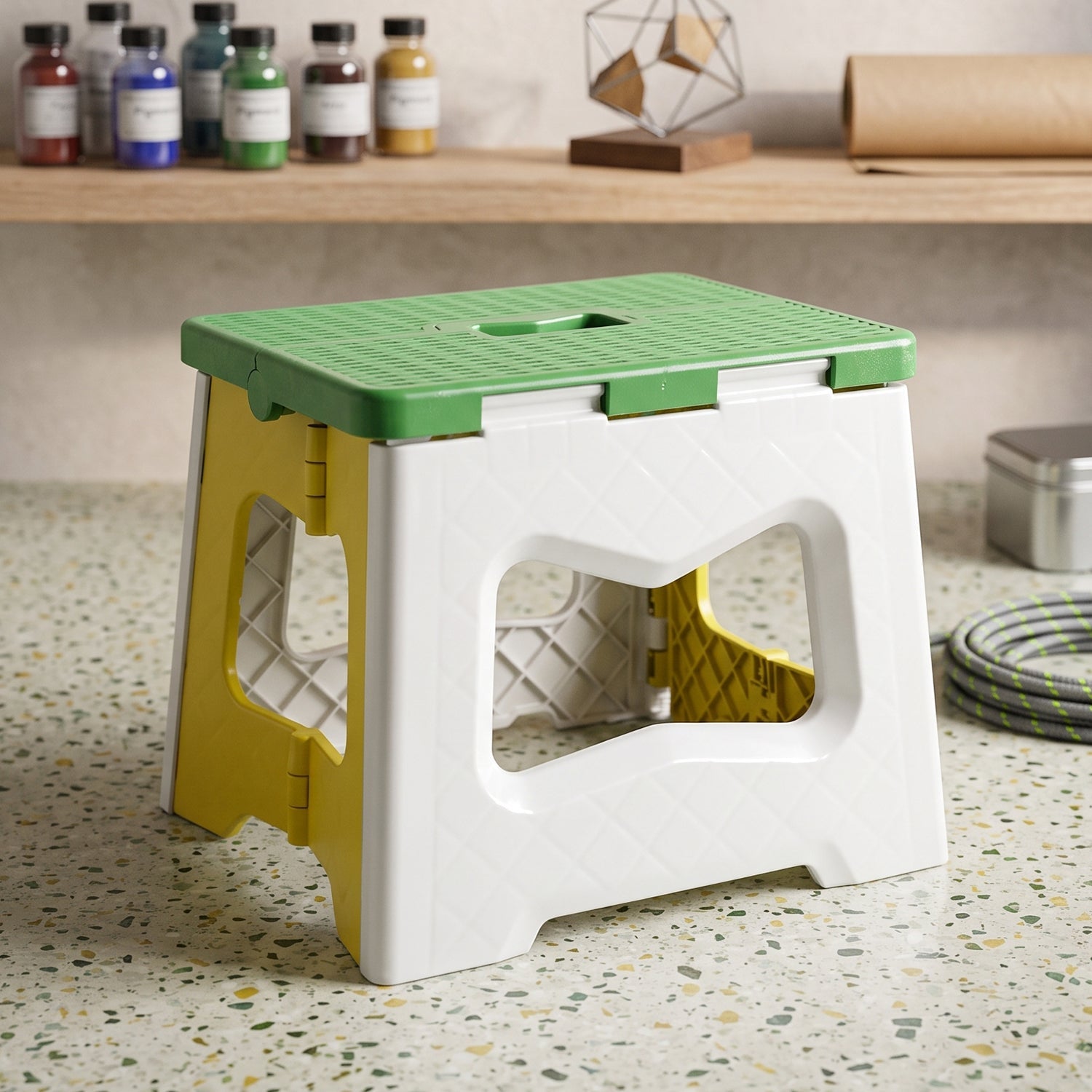 A green, white, and yellow plastic folding stool sits on a speckled counter with various craft supplies in the background.