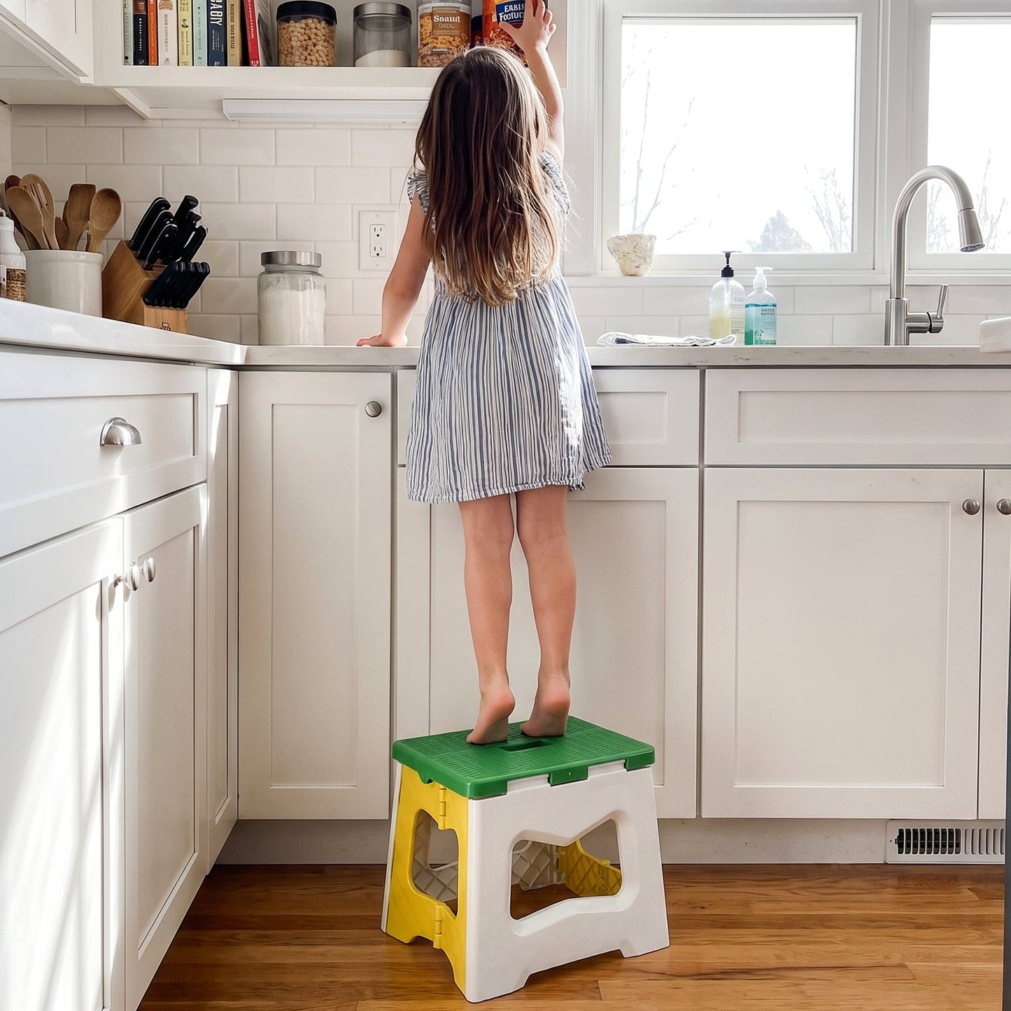 A child stands barefoot on the green, white, and yellow plastic stool, reaching for an item above a kitchen sink.