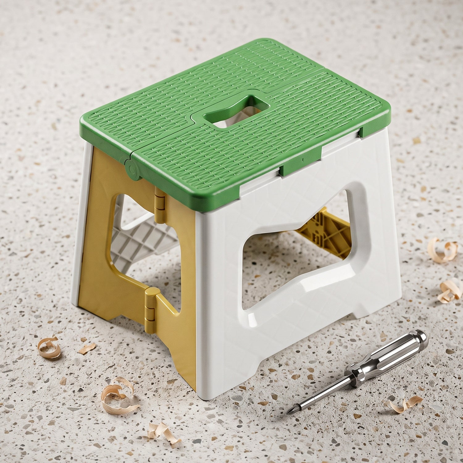 The green, white, and yellow plastic folding stool is on a speckled surface with wood shavings and a screwdriver nearby.