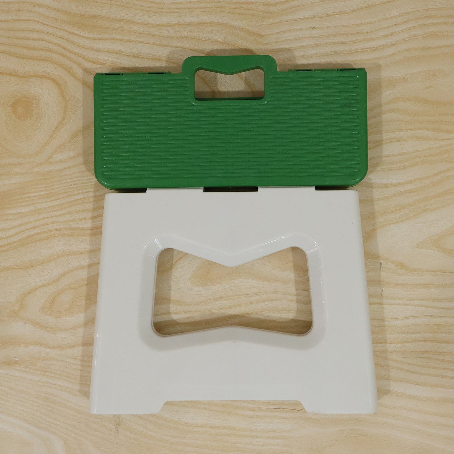 A green and white plastic folding stool is shown partially folded, revealing its carry handle, on a wooden surface.