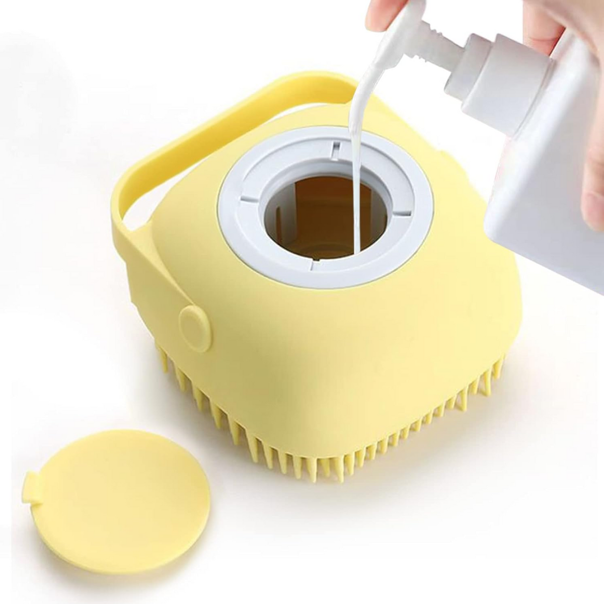 A hand pours white liquid soap from a bottle into the open top of a yellow silicone bath brush, with its cap beside it.