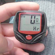 A hand holding a small black and red bicycle computer displaying 0 KM/hr and Hr.