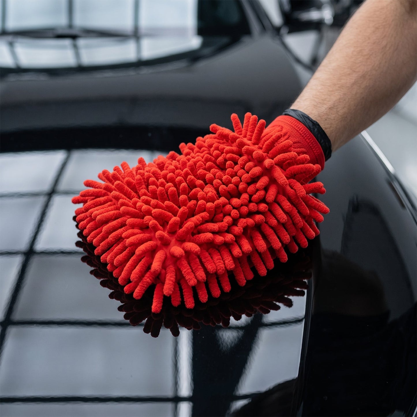 A hand wearing a red microfiber chenille wash glove cleans the shiny black hood of a car.