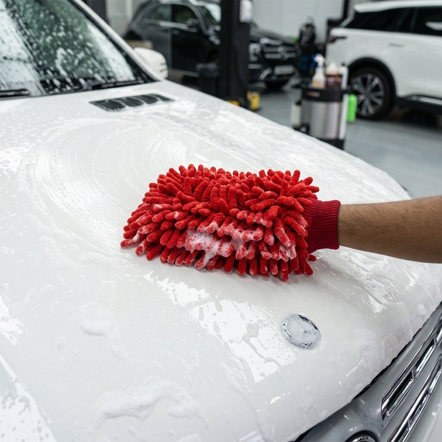 A hand wearing a red microfiber chenille wash glove cleans the sudsy white hood of a car in a garage setting.