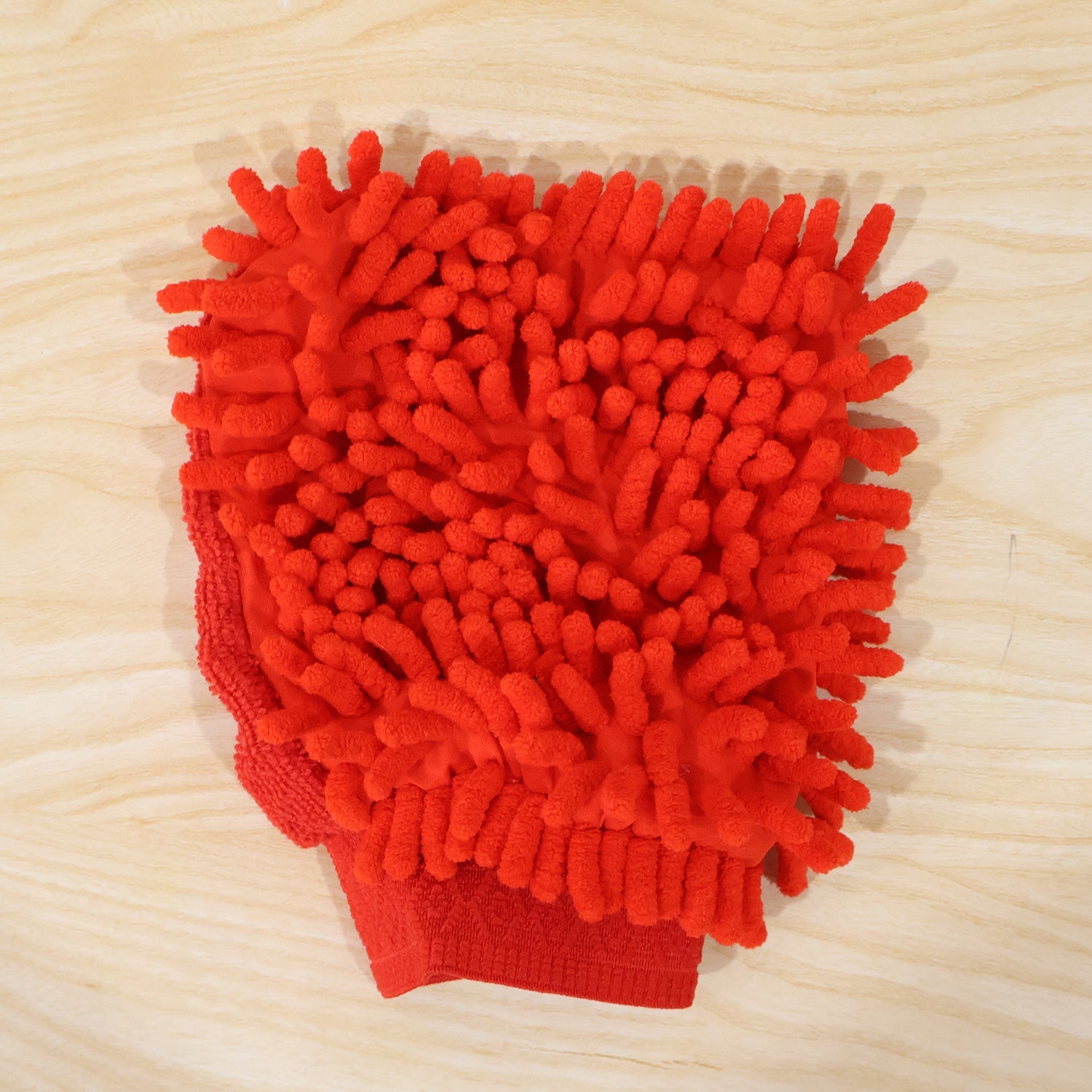 A red microfiber chenille wash glove with a textured surface lies flat on a light wooden surface.